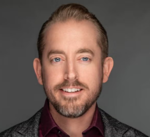 Man with light brown hair wearing a dark black and gold jacket over a maroon collared shirt.