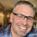 A man with short dark and gray hair wearing a blue, red and white plaid shirt and black glasses.