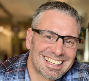 A man with short dark and gray hair wearing a blue, red and white plaid shirt and black glasses.