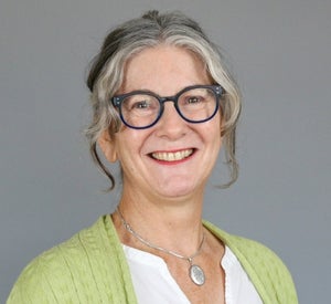 Woman with gray hair pulled back wearing dark gray glasses, a light green sweater over a white top and a silver pendant necklace.