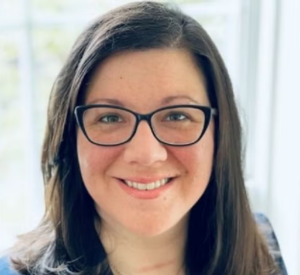 A woman with long brown hair and black glasses wearing a dark blue blazer.