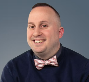 Man with very short brown hair wearing a a dark blue sweater and a light pink and gray bow tie.