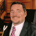 A man with short dark hair wearing a black suit jacket over a white collared shirt with a red and white striped tie.