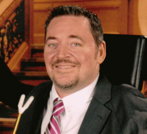 A man with short dark hair wearing a black suit jacket over a white collared shirt with a red and white striped tie.