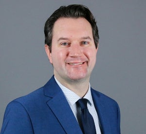 Man with short dark hair wearing a blue suite with a dark blue tie.