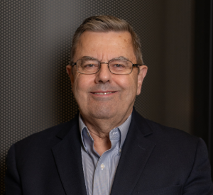 Man with short brown hair wearing a black suit jacket over a gray collared shirt and wire-rimmed glasses.