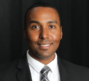 Man with short black hair wearing a black suit jacket over a white collared shirt with a gray and white striped tie.