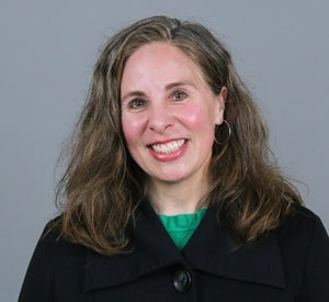 Woman with grey-blonde medium length hair, wearing a black sweater with a large button.