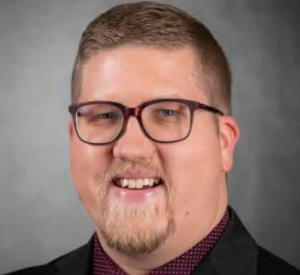 A man with short brown hair wearing glasses and a black suit jacket over a dark red collared shirt.