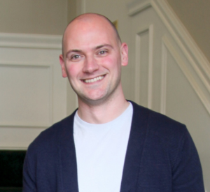 Bald man wearing a black cardigan over a white shirt.