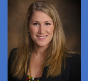 Woman with dark blonde hair wearing a black blazer over a patterend blouse.