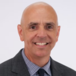 Bald man wearing a dark gray suit jacket over a gray and white collared shirt and a black tie.