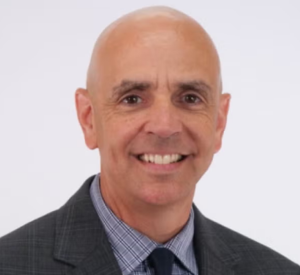 Bald man wearing a dark gray suit jacket over a gray and white collared shirt and a black tie.