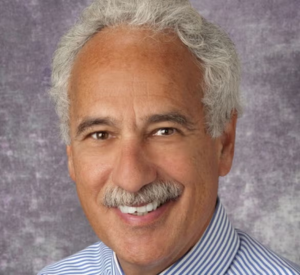 Man with white hair and a mustache wearing a white and gray striped collared shirt.