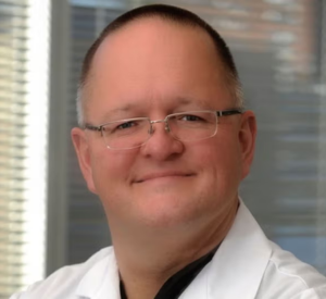 A man with very short brown and gray hair wearing a white lab coat over a black shirt.