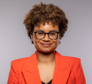 Woman with auburn curly hair wearing an orange blazer over a black shirt.