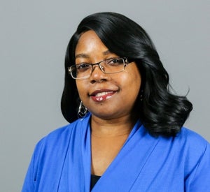 Woman with medium length, black hair wearing glasses, dangle earrings and a bright blue top.