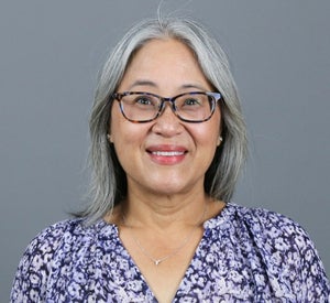 Woman with medium length grey hair wearing glasses and a purple floral top.