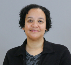 A woman with short, dark curly hair wearing a black cardigan over a black and gray patterned shirt.