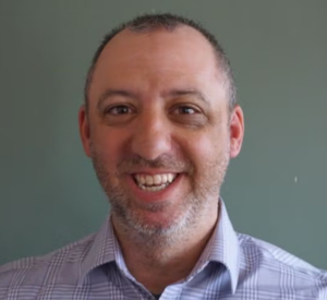 Man with very short dark hair and a gray beard wearing a gray collared shirt.