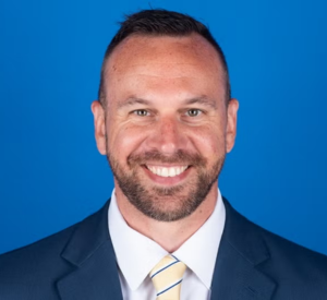 Man with short dark hair wearing a dark blue suit jacket over a white collared shirt with a light yellow and blue striped tie.