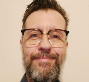 Man with dark hair and beard wearing black and clear glasses wearing a black collared shirt.