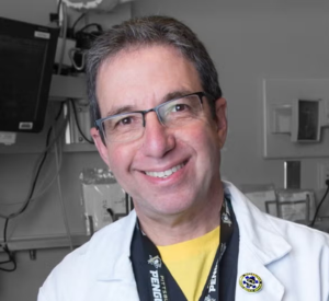 Man with dark and gray hair wearing a white lab coat over a yellow shirt.