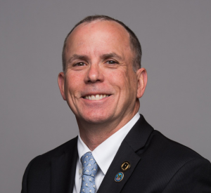 Man with very short brown hair wearing a black suit jacket over a white collared shirt and a light blue tie.