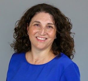 Woman with medium length, dark brown, curly hair wearing a bright blue top.