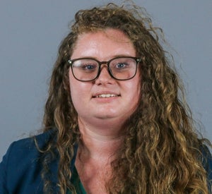 Woman with long, dark blonde, curly hair, wearing glasses and a dark blue blazer.