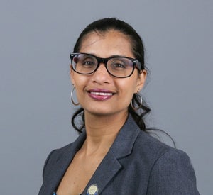 Woman with black hair pulled back in a ponytail wearing bold glasses, hoop earrings and a dark grey blazer.