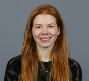 Woman with long red hair wearing a black and brown patterned shirt.