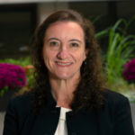 Woman with dark brown curly hair wearing a black blazer over a white blouse.