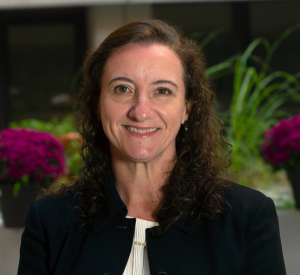 Woman with dark brown curly hair wearing a black blazer over a white blouse.