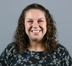 Woman with medium length, dark brown, curly hair wearing a black and white floral pattern shirt.