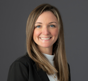 Woman with dark blonde hair wearing a black blazer over a white turtleneck and gold necklaces.