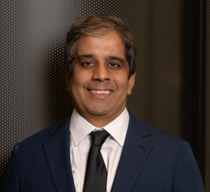 Man with short brown and gray hair wearing a black suit jacket over a white collared shirt with a black tie.