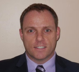 Man with short auburn hair wearing a black suit jacket over a light purple collared shirt and a dark purple tie.