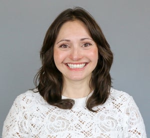 Woman with medium length, dark brown hair wearing a white floral cutout shirt.