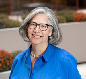 Woman with short gray hair wearing a collared bright blue blouse, a white beaded necklace and brown glasses.