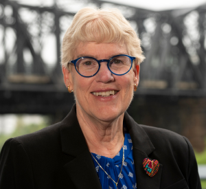 Woman with short white blonde hair wearing a black suit jacket over a dark blue patterned blouse with a red heart pin and blue glasses.