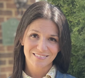 A woman with dark brown hair wearing a denim jacket over a yellow floral collared shirt.