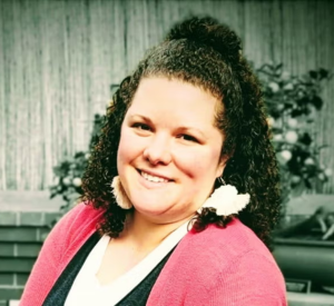 Woman with black curly hair wearing a pink cardigan over a black and white blouse.