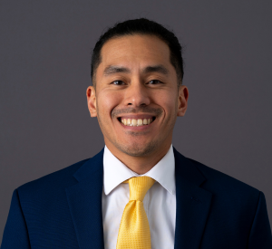 Man with short dark hair wearing a black suit jacket over a white collared shirt and a yellow tie.