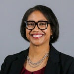 Woman with short dark hair wearing bold glasses, a silver statement necklace and a black blazer.