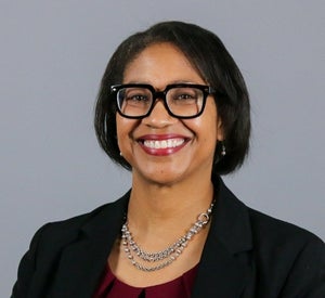 Woman with short dark hair wearing bold glasses, a silver statement necklace and a black blazer.