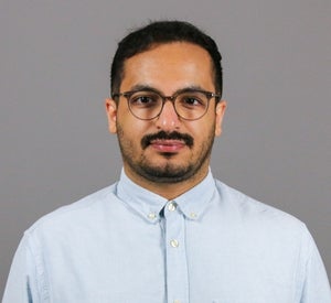 Man with dark hair, mustache and light beard wearing a light blue button up shirt.