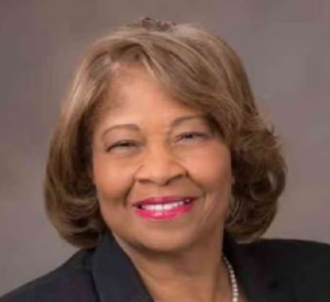 A woman with short brown hair wearing a black blazer and a pearl necklace.