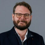 Man with short brown hair and beard. Wearing glasses and a black jacket.