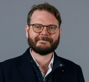 Man with short brown hair and beard. Wearing glasses and a black jacket.